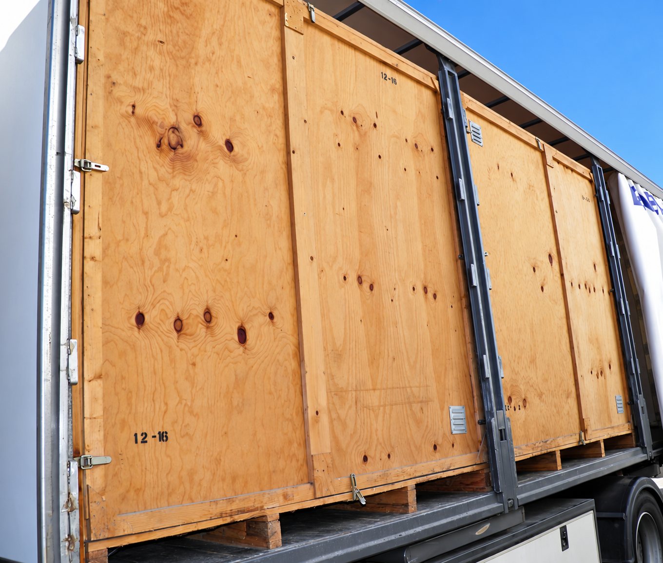 Plusieurs grandes caisses en contreplaqué posées sur des palettes à l'intérieur d'une remorque de camion à bâche coulissante, sous un ciel bleu.