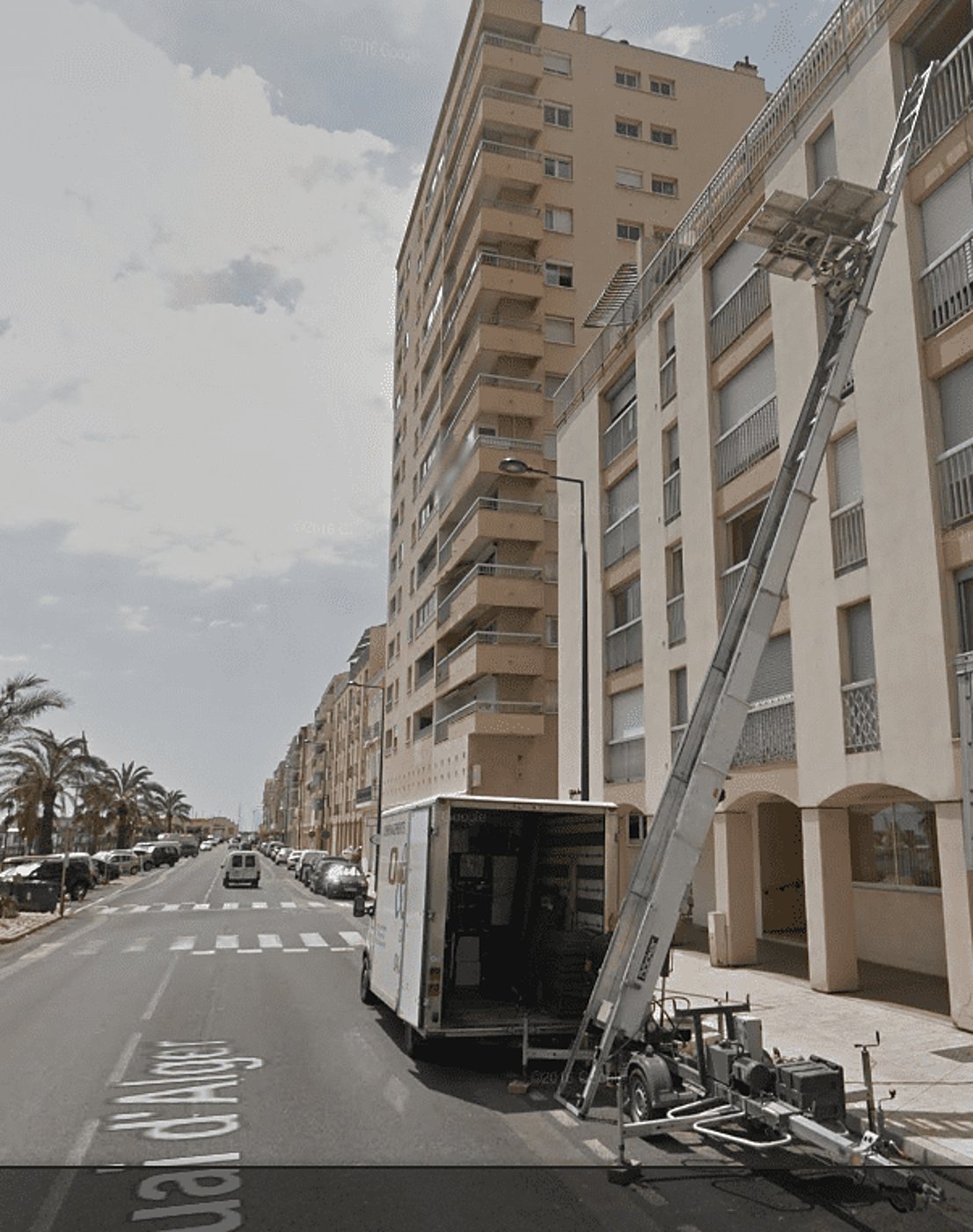 Un camion de déménagement stationné dans une rue bordée de palmiers dresse un monte-meubles incliné contre la façade d'un immeuble résidentiel.