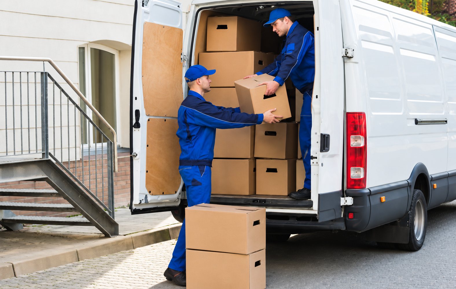 Deux déménageurs en uniforme bleu et casquette sortent des cartons de l'arrière d'une camionnette blanche garée devant un immeuble.