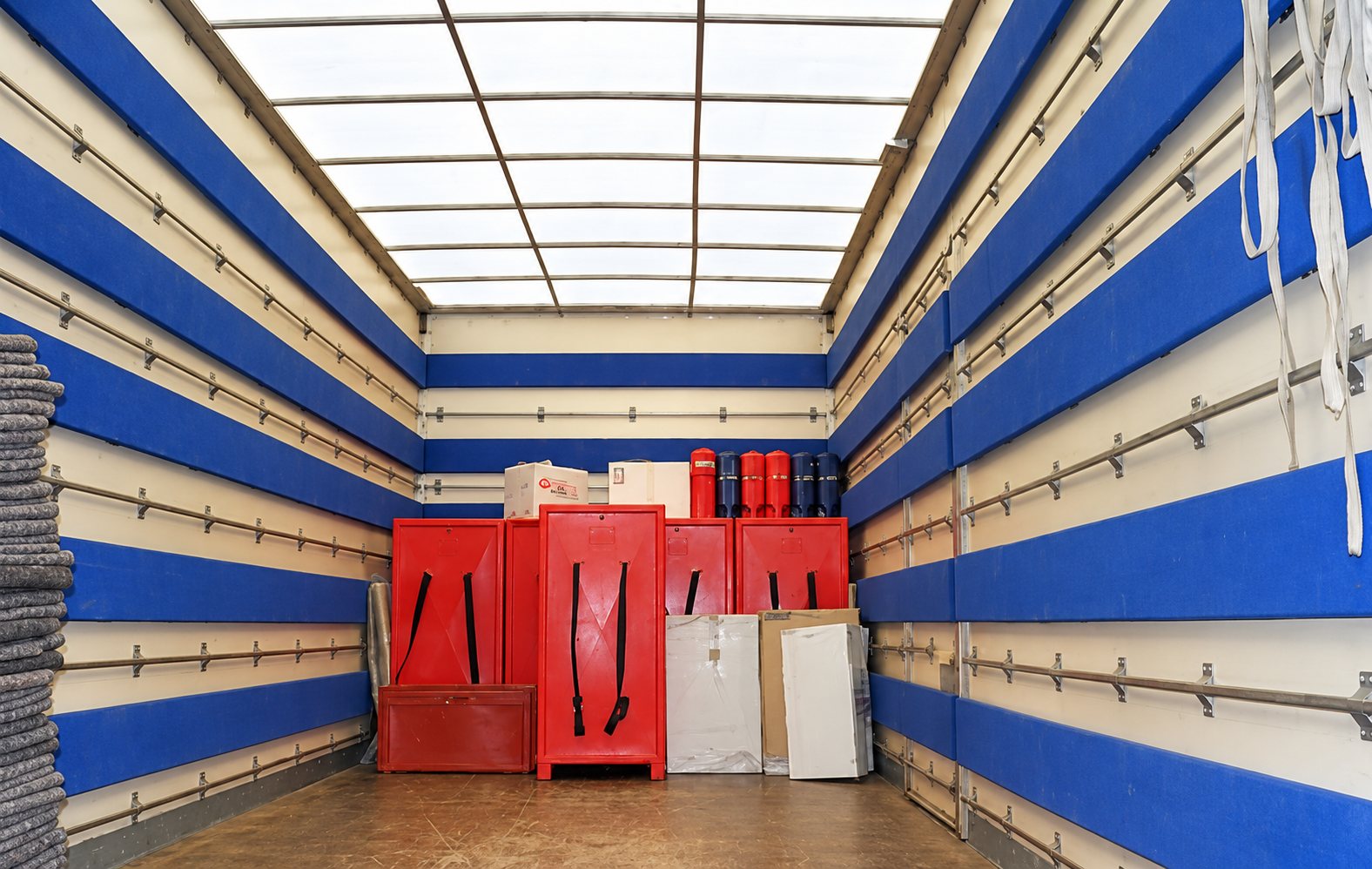 Vue de l'intérieur d'un camion de déménagement aux parois matelassées bleues et blanches, contenant des armoires de transport rouges, des cartons et des panneaux.