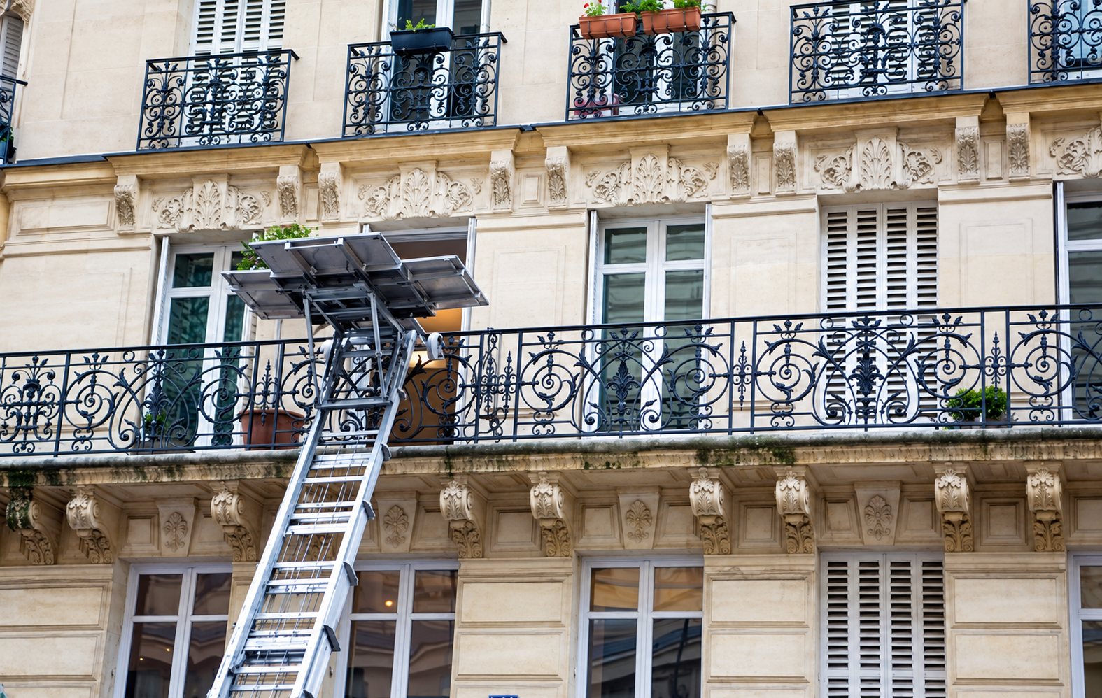 Un monte-meubles à échelle est déployé le long de la façade d'un immeuble parisien en pierre claire, jusqu'à un balcon à garde-corps en fer forgé.