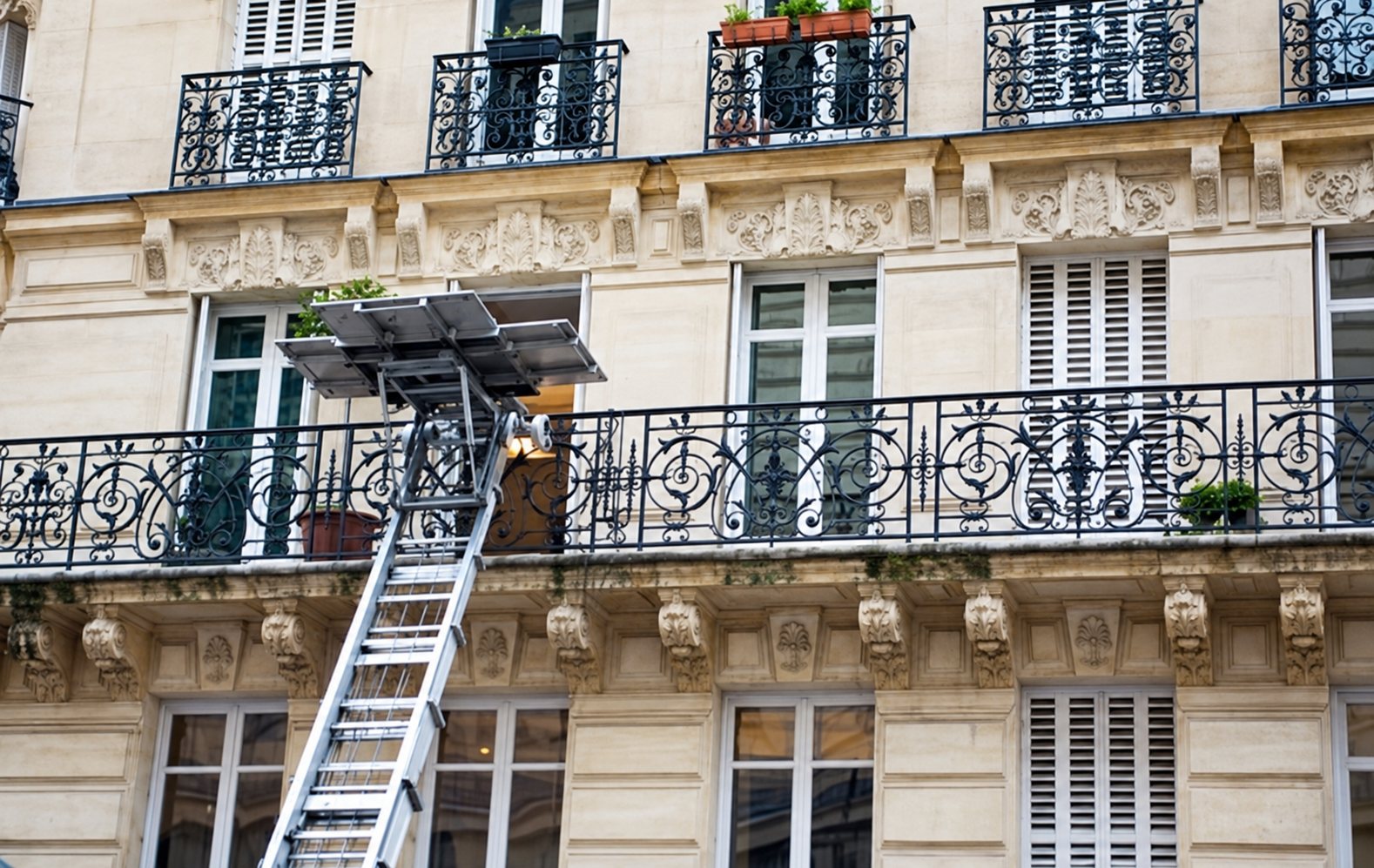 Un monte-meubles avec son échelle est dressé contre la façade en pierre d'un immeuble ancien aux balcons en fer forgé.